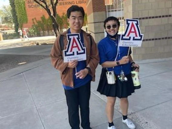 Two people hold the UArizona "A" in front of Koffler building