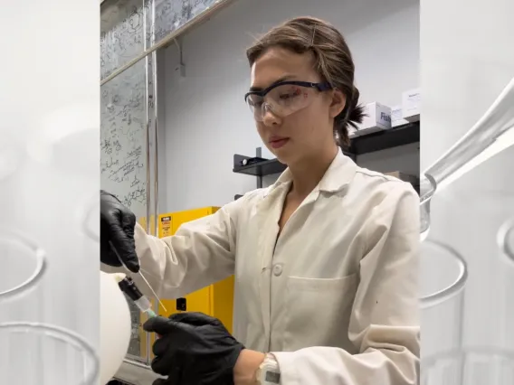 Asia Richardson looking down, holding a syringe attached to a white balloon, and in a lab wearing a lab coat and safety goggles.