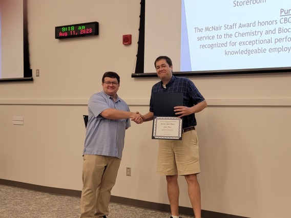 Justan Klaus (right) receiving the Dr. Harold McNair Staff Award from Craig Aspinwall (left). Both standing in front of a projector screen and a wall clock on the top left.