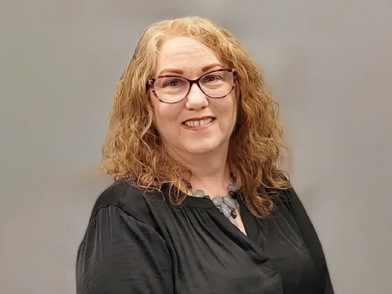 Nancy Louks smiling towards the cameras, standing in front of a gray background, wearing glasses and a black blouse.