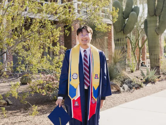 Andrew Leung standing outside Old Main wearing a cap and gown.