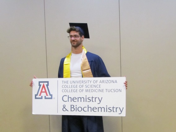 Devin Collins wearing a graduation cap and gown holding a UofA Chemistry and Biochemistry Sign