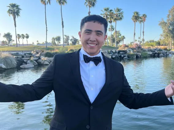 Noah Canisales, facing the camera wearing a black suit and tie. Standing in front of a lake and a row of palm trees.