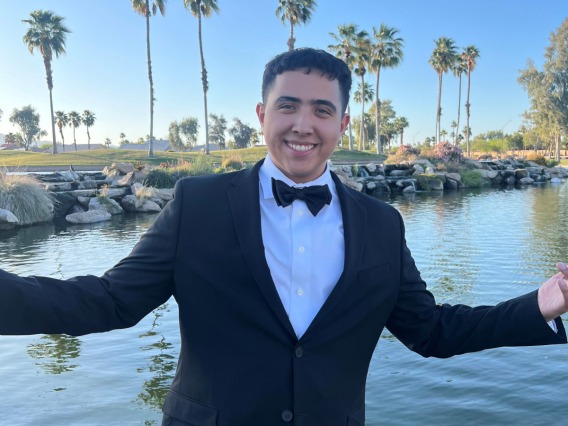 Noah Canisales, facing the camera wearing a black suit and tie. Standing in front of a lake and a row of palm trees.