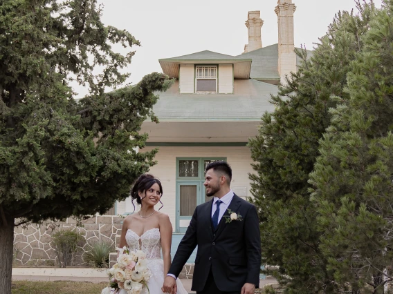 Aurora Hurtado and Ramon Cano in wedding attire, standing between a line of trees and a small building