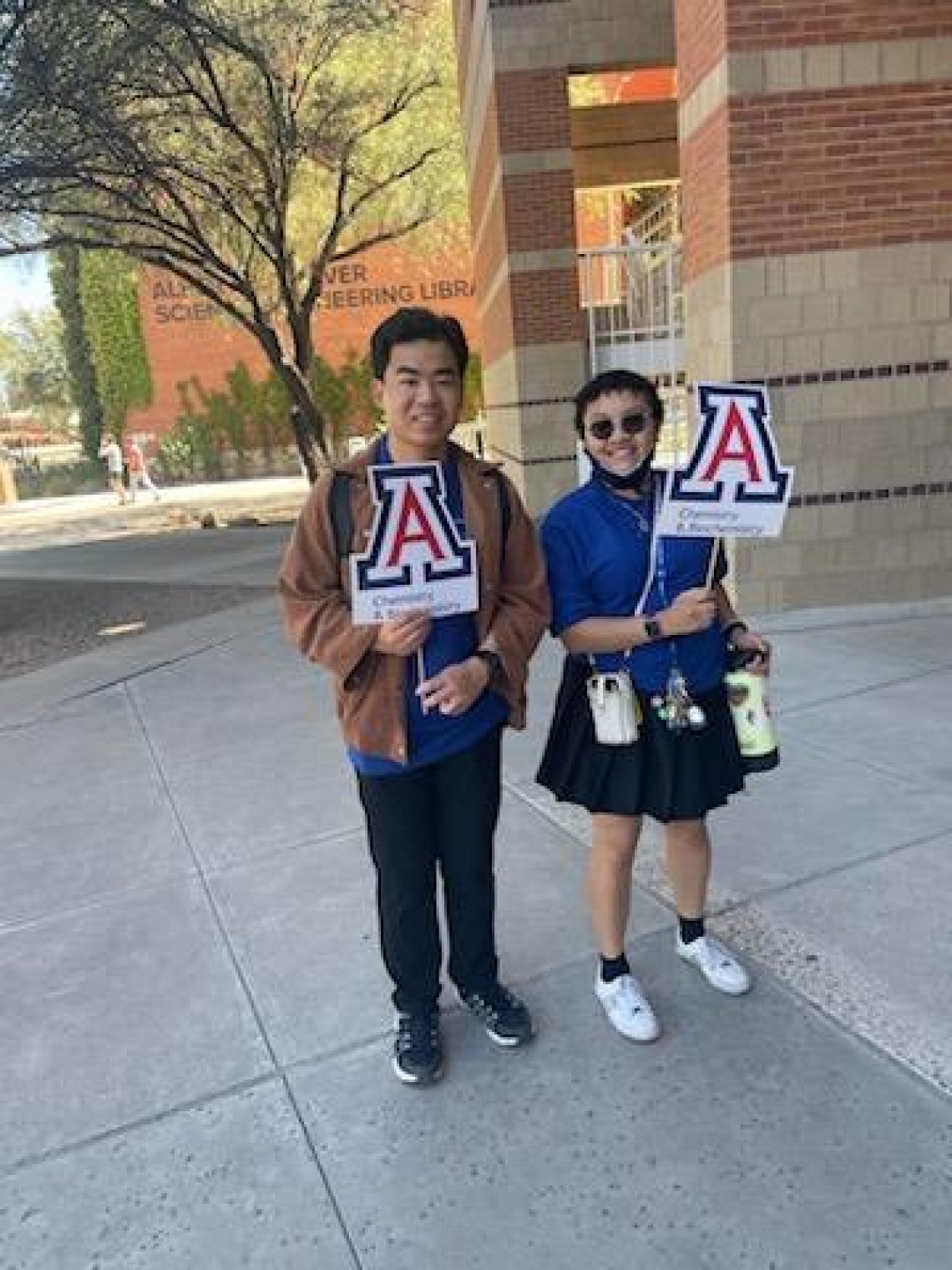 Two people hold the UArizona "A" in front of Koffler building