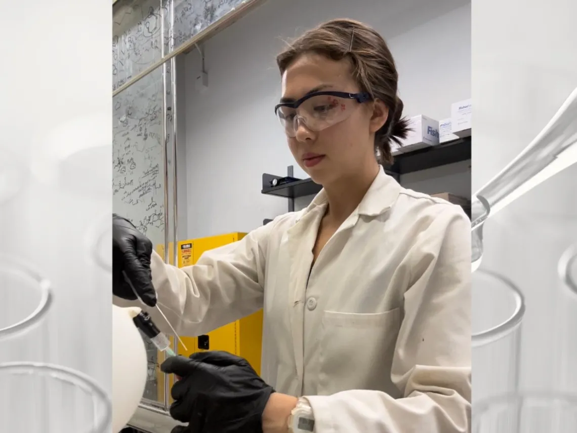 Asia Richardson looking down, holding a syringe attached to a white balloon, and in a lab wearing a lab coat and safety goggles.