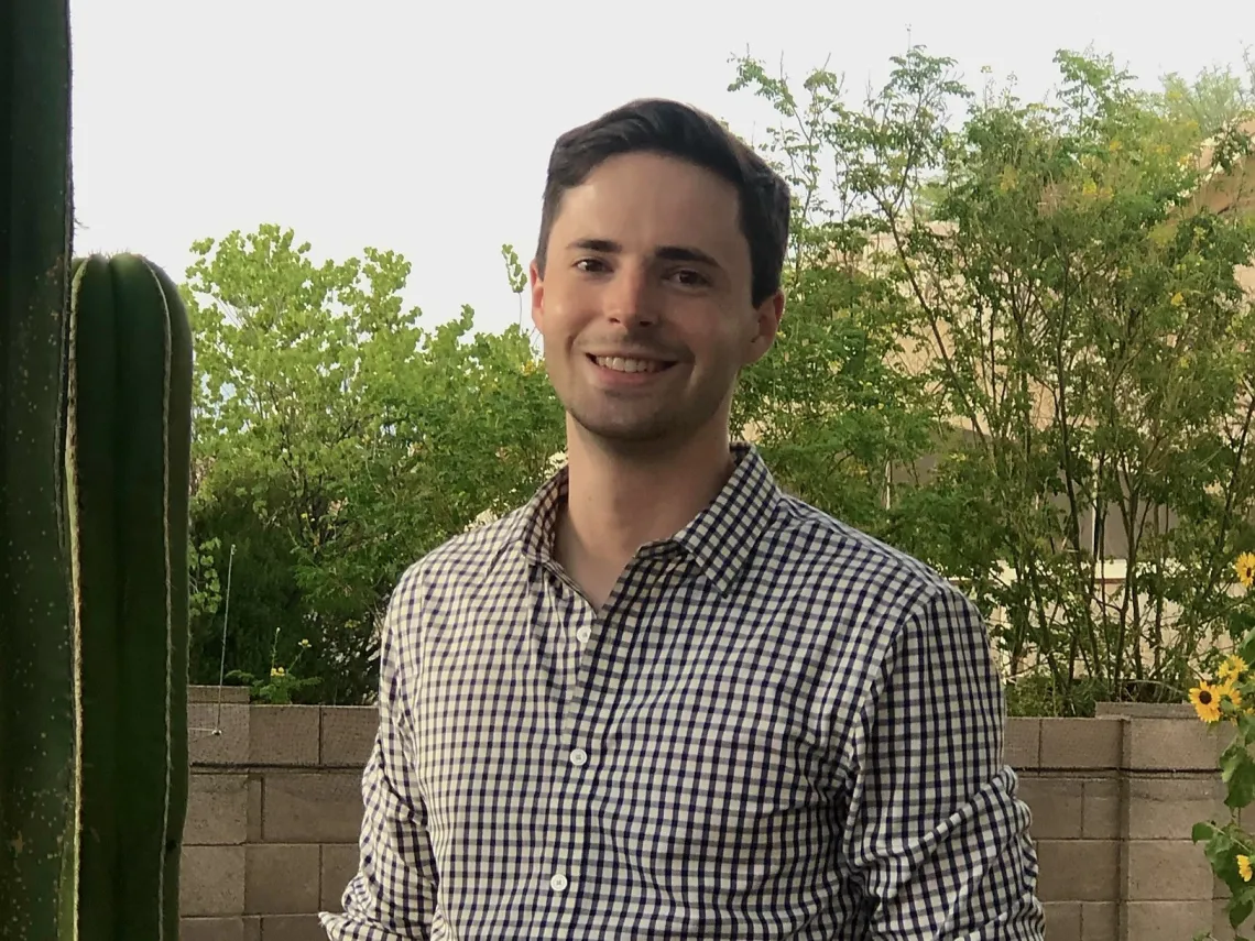 Christopher Marshall smiling towards the camera, wearing a plaid shirt and standing in front of a cactus on the left side.