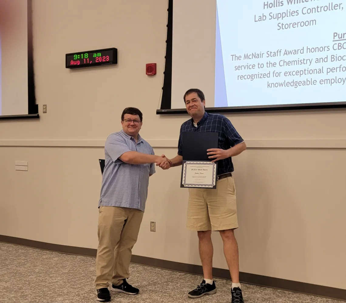 Justan Klaus (right) receiving the Dr. Harold McNair Staff Award from Craig Aspinwall (left). Both standing in front of a projector screen and a wall clock on the top left.