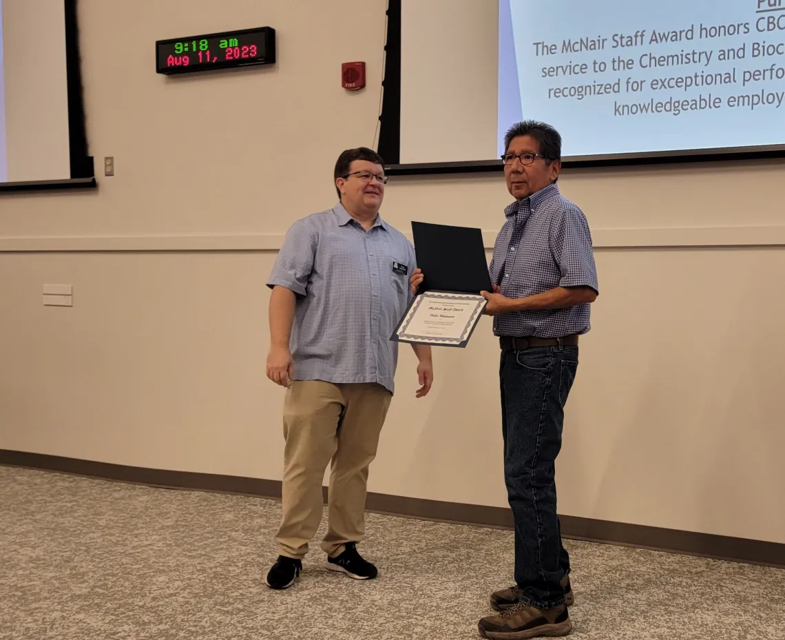 Hollis Whitewater (right) receiving the Dr. Harold McNair Staff Award from Craig Aspinwall (left). Both standing in front of a projector screen and a wall clock on the top left.