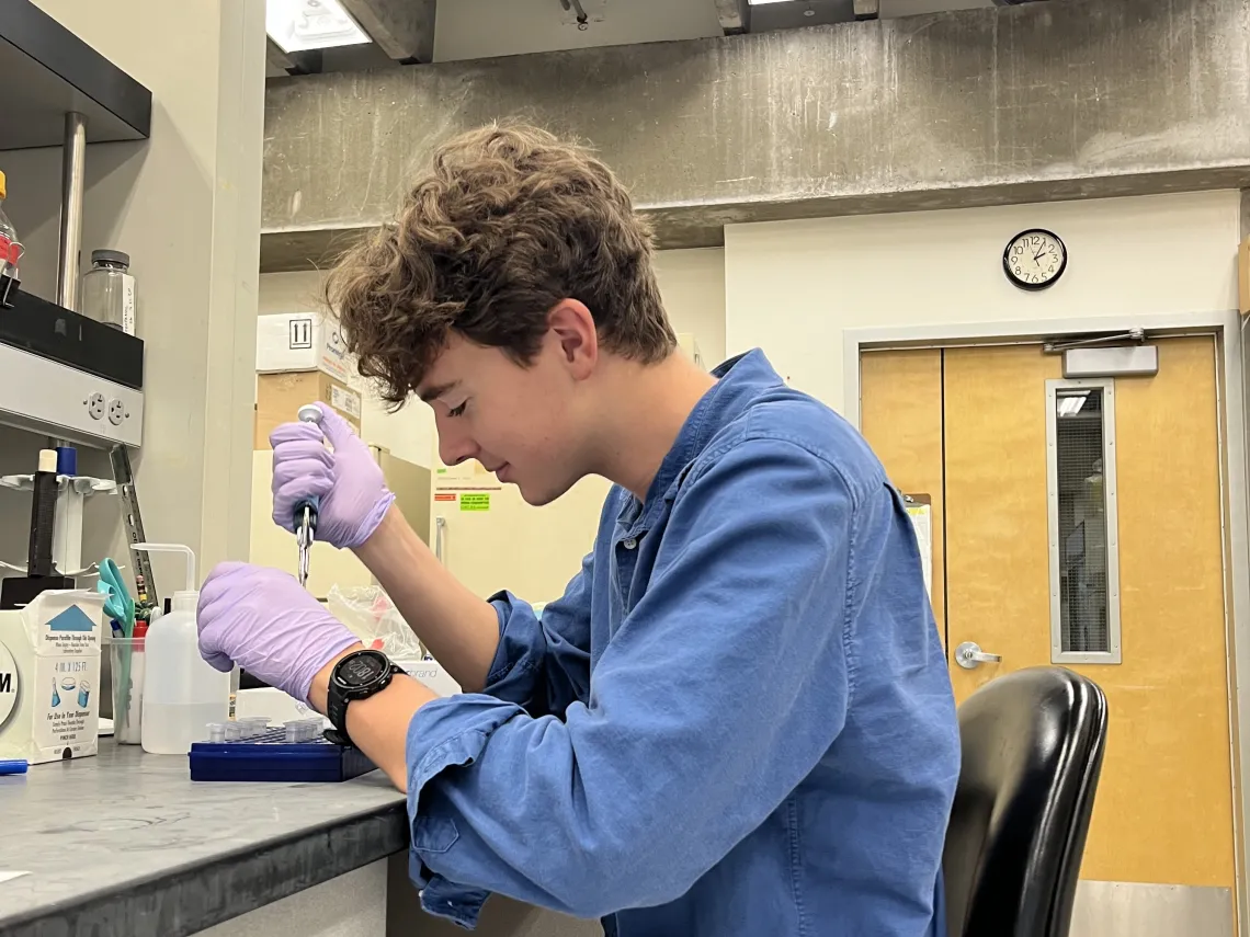 Jackson Taylor sitting on a chair in a lab, facing the left wearing purple surgical gloves holding testing tubes.