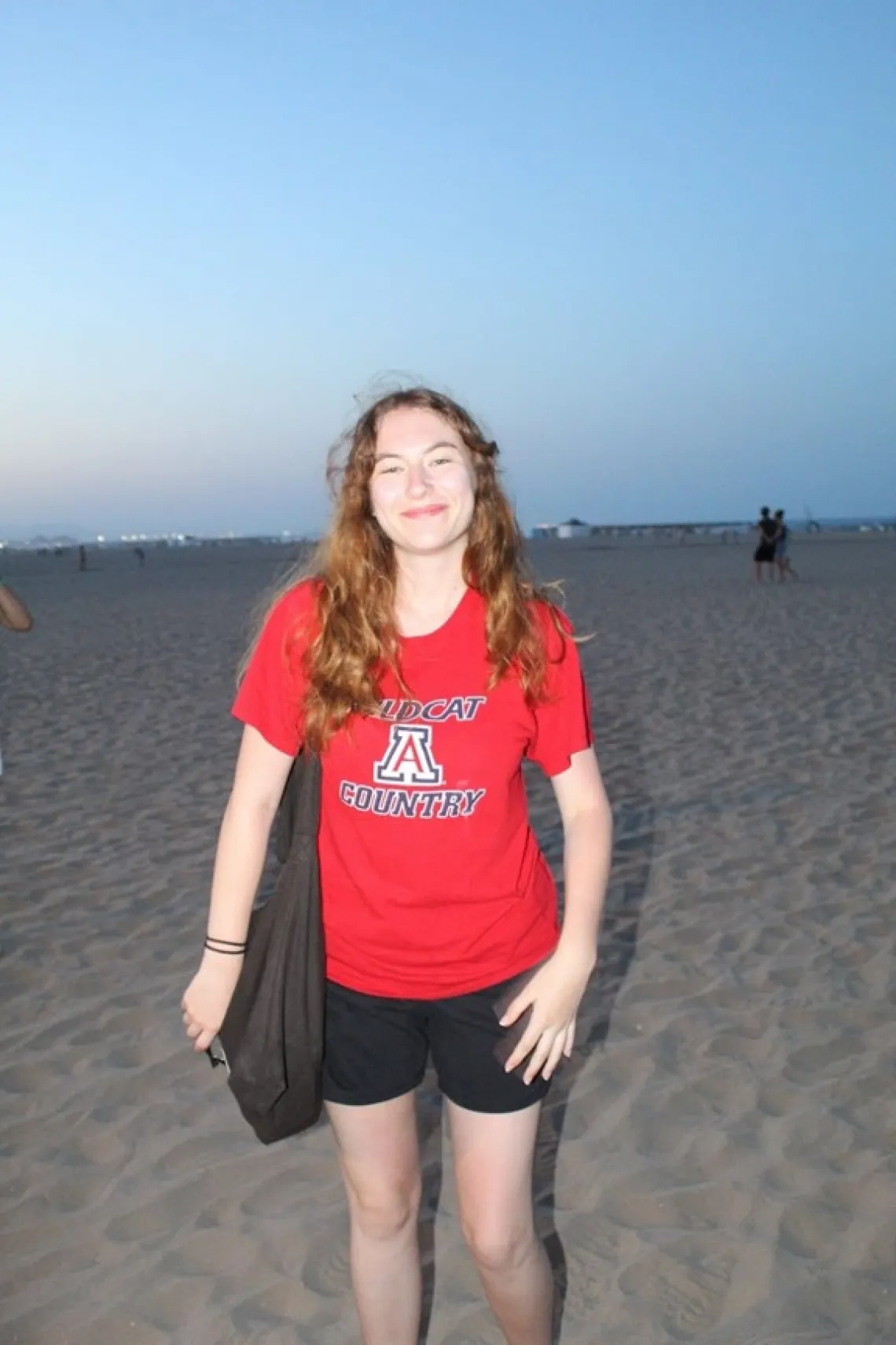 Olivia Seagraves at the beach, smiling towards the camera wearing a red University of Arizona shirt and a black tote bag.