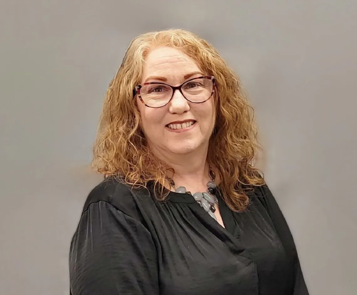 Nancy Louks smiling towards the cameras, standing in front of a gray background, wearing glasses and a black blouse.