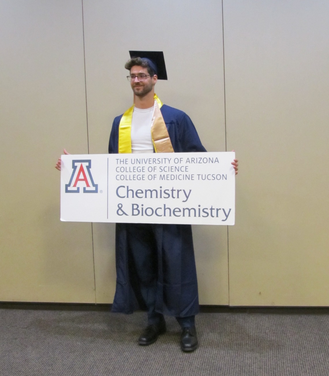Devin Collins wearing a graduation cap and gown holding a UofA Chemistry and Biochemistry Sign