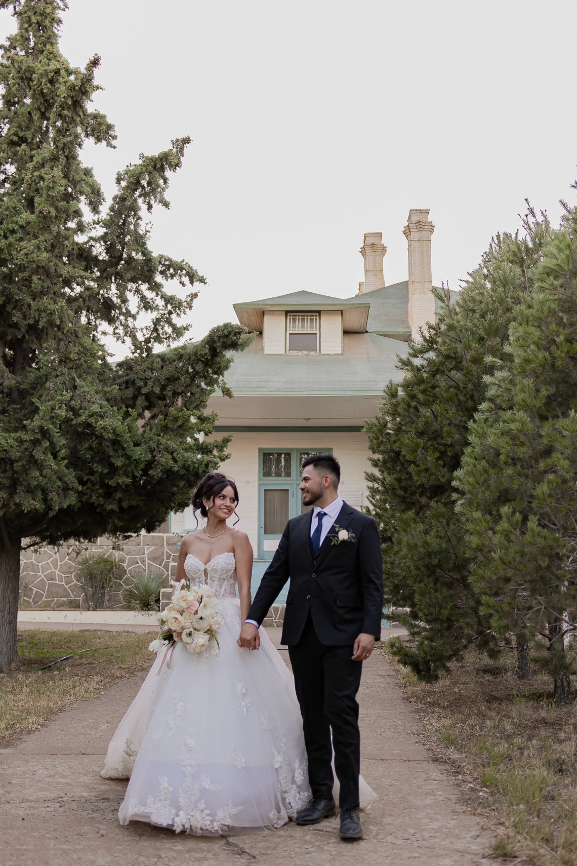 Aurora Hurtado and Ramon Cano in wedding attire, standing between a line of trees and a small building